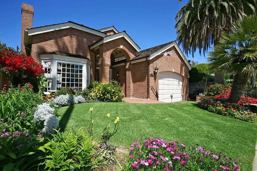 The funded La Jolla residence: a two-story brick house with a chimney, bay window, arched entry and attached garage, with a lawn, flower beds and a palm in front.