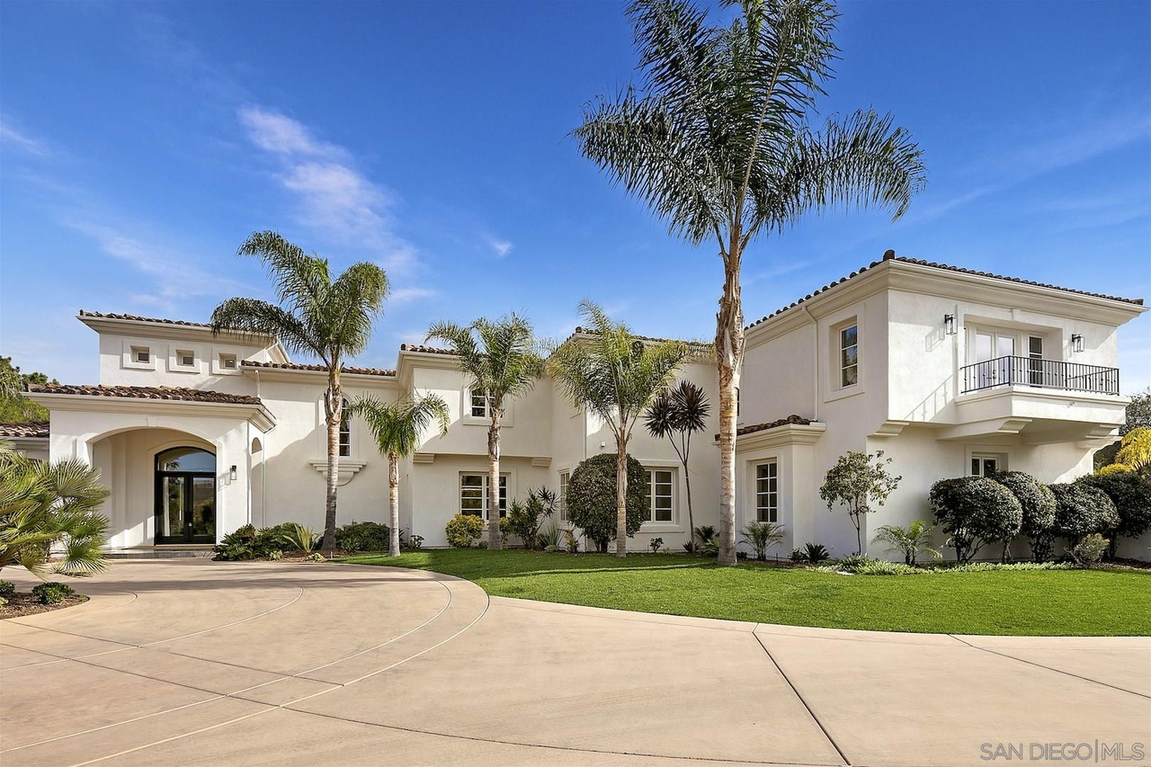 The funded Rancho Santa Fe residence: a white two-story home with a tile roof, arched entry and balcony, tall palms and a lawn beside a circular concrete drive.